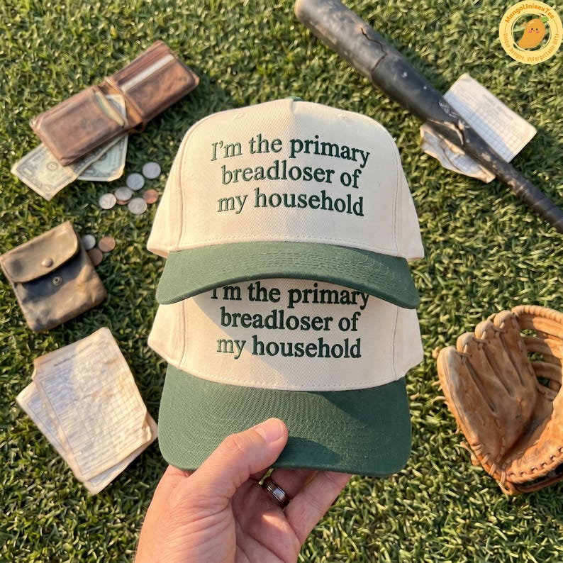 K&ouml;nnte beinhalten: Zwei Baseballkappen mit cremefarbenem Oberteil und gr&uuml;nem Schirm sind &uuml;bereinander gestapelt. Der Text "I'm the primary breadloser of my household" ist in Gr&uuml;n auf die Vorderseite jeder Kappe gedruckt. Ein Baseballschl&auml;ger, ein Handschuh und eine Geldb&ouml;rse sind im Hintergrund.