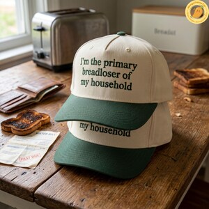 K&ouml;nnte beinhalten: Zwei beige Baseballkappen mit dunkelgr&uuml;nen Schirmen sind auf einem Holztisch gestapelt. Auf der oberen Kappe steht in dunkelgr&uuml;ner Schrift "I'm the primary breadloser of my household". Auf der unteren Kappe steht "my household".