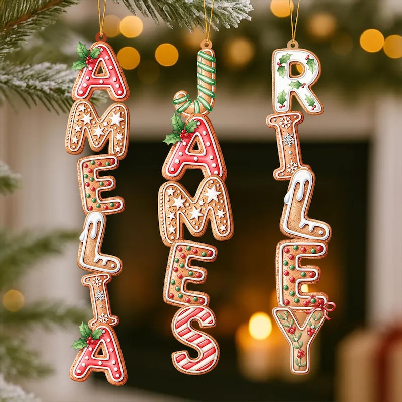 May include: Christmas ornaments spelling out the names "Amelia" and "James Riley" in gingerbread cookie letters. The letters are decorated with red, white, and green icing, holly, and star-shaped sprinkles. The ornaments hang from gold strings.