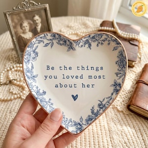 May include: Heart-shaped ceramic dish with a blue floral design and gold trim. The dish features the text "Be the things you loved most about her" and a small blue heart. Decorative item.
