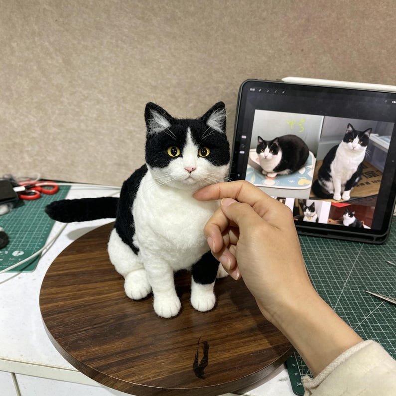 Op de afbeelding: Een naaldgevilte kattenfiguur met een zwart-witte vacht, puntige oren en amberkleurige ogen. De kat is afgebeeld in een zittende positie met de staart naar achteren gestrekt, met een realistische textuur en gedetailleerde gelaatstrekken.
