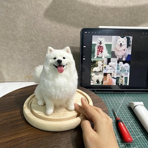 Op de afbeelding: Een naaldgevilte witte Samojeed hondfiguur met een zwarte neus en roze tong, zittend op een ronde houten basis. Daarnaast toont een tablet een collage van foto's van een witte hond, samen met een gereedschap met rood handvat en een wit apparaat.