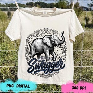 May include: A cream-colored short-sleeve t-shirt featuring a black and white graphic illustration of an elephant. The elephant is centered within an ornate, swirling circular frame. Below the elephant, the word Swagger is written in a bold script.
