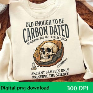 Op de afbeelding: Een crèmekleurig sweatshirt met een grafische print met de tekst OLD ENOUGH TO BE CARBON DATED, DETERMINE THE AGE, COLLECT DATA, ANCIENT SAMPLES ONLY, PRESERVE THE SCIENCE, een menselijke schedel op een rots, een kolf en een pincet.
