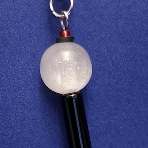 May include: A keychain charm featuring a silver-toned lobster clasp attached to a small red bead, a translucent white spherical bead with internal silver glitter, and a smooth, cylindrical black bead, finished with a small silver-toned metal ring.