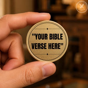 Peut inclure: Un jeton circulaire en métal doré à la surface texturée. Le centre présente le texte noir "YOUR BIBLE VERSE HERE" encadré par deux lignes horizontales, chacune accentuée par une petite étoile à quatre branches au-dessus et en dessous du texte.