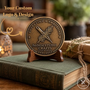 May include: A circular bronze-toned medallion featuring an embossed design of a crossed pen and leafy branch. The text reads ENGRAVING & EMBOSSING, habitat STUDIO CRAFTS, and EST. 2025. The medallion is displayed on a small wooden stand.