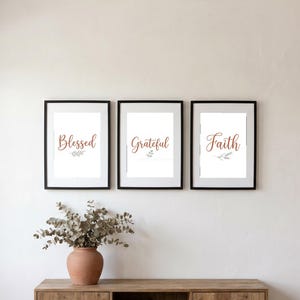 Könnte beinhalten: Drei rechteckige Wandbilder in schwarzen Rahmen. Jeder weiße Druck zeigt braune Schreibschrift mit den Wörtern Blessed, Grateful und Faith, ergänzt durch kleine graue botanische Illustrationen unter jedem Wort an einer schlichten Wand.