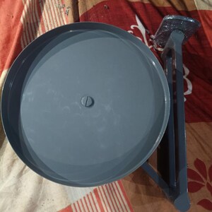 May include: A grey metal circular platform with a central screw, attached to a vertical support arm and a rectangular base plate. The structure features a smooth, uniform grey finish throughout, with a flat, round top surface and a sturdy, angular frame.