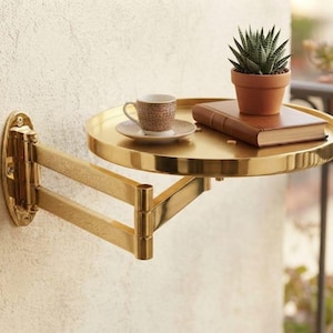 May include: A gold-toned metal wall-mounted swing arm shelf with a circular tray. The shelf holds a small brown patterned ceramic espresso cup on a matching saucer, a closed brown hardcover book, and a small green succulent in a terracotta pot.