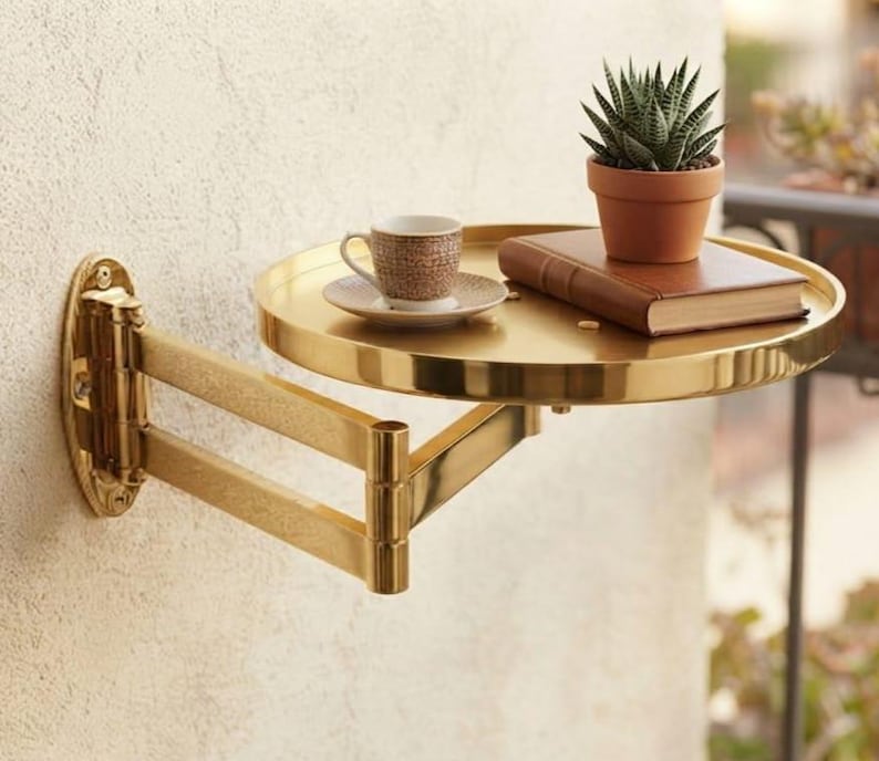 May include: A gold-toned metal wall-mounted swing arm shelf with a circular tray. The shelf holds a small brown patterned ceramic espresso cup on a matching saucer, a closed brown hardcover book, and a small green succulent in a terracotta pot.
