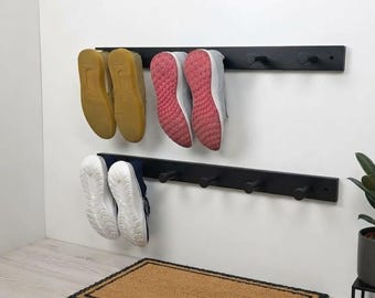 May include: Two horizontal black wall-mounted racks with cylindrical pegs for shoe storage. The top rack holds a pair of tan trainers and a pair of grey trainers with pink soles. The bottom rack holds a pair of white trainers with blue accents.