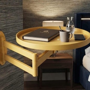 May include: A mustard yellow wall-mounted swing-arm side table with a circular tray top. The table features a hinged, multi-jointed arm attached to a vertical oval wall plate. A black book, a grey mug, a glass carafe, and a smartphone sit on top.