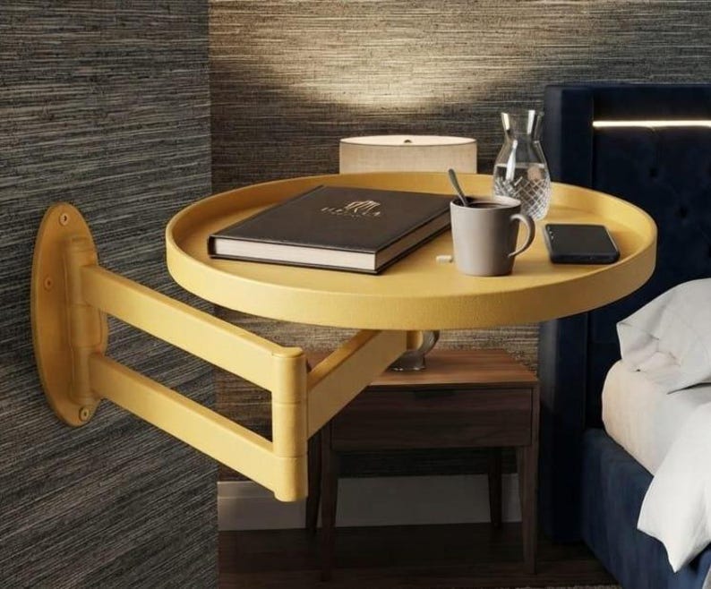 May include: A mustard yellow wall-mounted swing-arm side table with a circular tray top. The table features a hinged, multi-jointed arm attached to a vertical oval wall plate. A black book, a grey mug, a glass carafe, and a smartphone sit on top.