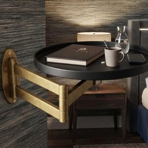 May include: A wall-mounted, round black tray table with a brass-coloured, multi-jointed articulating arm. The tray holds a closed dark book, a light-coloured ceramic mug, a clear glass carafe, and a black smartphone on its surface.
