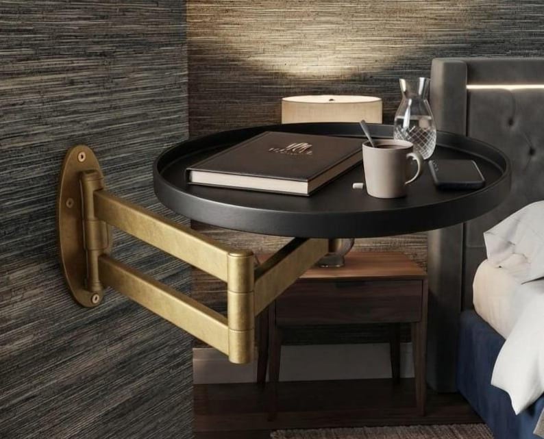 May include: A wall-mounted, round black tray table with a brass-coloured, multi-jointed articulating arm. The tray holds a closed dark book, a light-coloured ceramic mug, a clear glass carafe, and a black smartphone on its surface.