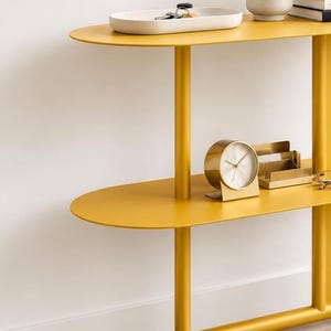 May include: A mustard yellow metal console table with two oval shelves and a curved base. The top shelf holds a white tray with keys and a potted succulent. The lower shelf features a gold-toned circular desk clock and a matching card holder.