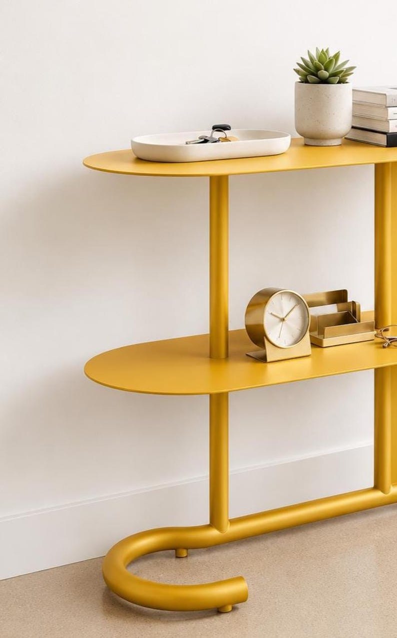 May include: A mustard yellow metal console table with two oval shelves and a curved base. The top shelf holds a white tray with keys and a potted succulent. The lower shelf features a gold-toned circular desk clock and a matching card holder.