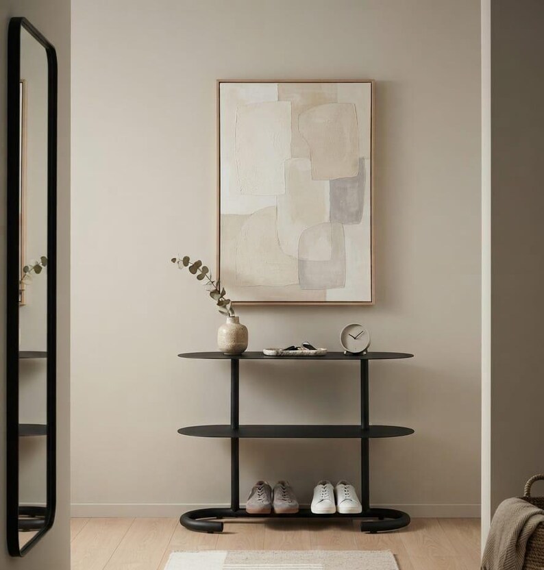 May include: A black metal console table with two oval shelves, featuring a light-coloured abstract painting in a wooden frame above it. On the top shelf sits a small vase with eucalyptus stems, a decorative tray, and a round analogue clock.