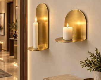 May include: Two wall-mounted candle holders made of brushed gold metal. Each holder features a vertical arched backplate and a small circular shelf. One holder supports a tall, slender white taper candle, while the other holds a thick white pillar candle.