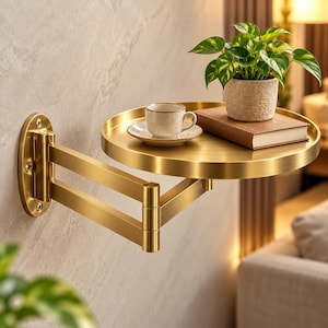 May include: A gold-toned metal wall-mounted swing arm shelf with a circular tray. The shelf holds a small white ceramic cup on a matching saucer, a closed brown book, and a potted green plant in a textured light-coloured container.
