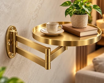 May include: A wall-mounted, brushed gold metal shelf with a circular tray and an adjustable, multi-jointed swing arm. The shelf holds a small ceramic cup on a saucer, a closed book with a brown cover, and a potted green plant in a textured pot.