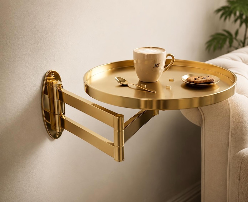 May include: A gold-toned, circular wall-mounted side table with an adjustable, multi-jointed swing arm. The round tray surface holds a beige ceramic mug with a logo, a small gold spoon, and a small plate with a single cookie.