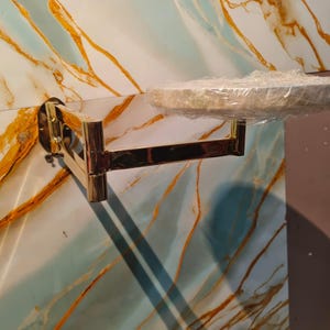 May include: A gold-toned, wall-mounted, articulated swing-arm bracket featuring a circular platform at the end. The platform is covered in a layer of clear plastic film. The metal arm has multiple hinged joints allowing for horizontal movement.