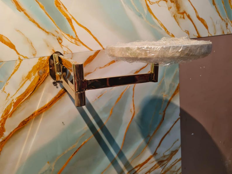 May include: A gold-toned, wall-mounted, articulated swing-arm bracket featuring a circular platform at the end. The platform is covered in a layer of clear plastic film. The metal arm has multiple hinged joints allowing for horizontal movement.