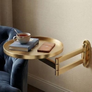 May include: A gold-toned, circular wall-mounted side table with an adjustable, multi-jointed swing arm. The flat, round surface holds a blue hardcover book, a white ceramic mug filled with tea, and a rectangular smartphone.
