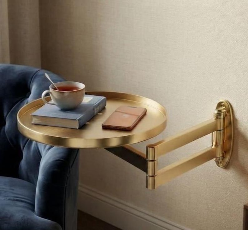 May include: A gold-toned, circular wall-mounted side table with an adjustable, multi-jointed swing arm. The flat, round surface holds a blue hardcover book, a white ceramic mug filled with tea, and a rectangular smartphone.