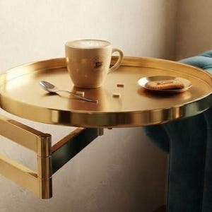 May include: A round gold-toned metal tray table with an adjustable swing arm. The tray holds a beige ceramic mug with latte art, a small silver spoon, two white sugar cubes, and a small plate with a single biscuit on it.