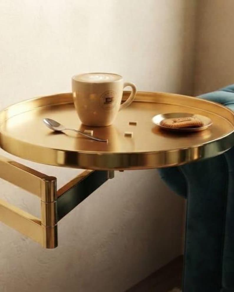 May include: A round gold-toned metal tray table with an adjustable swing arm. The tray holds a beige ceramic mug with latte art, a small silver spoon, two white sugar cubes, and a small plate with a single biscuit on it.