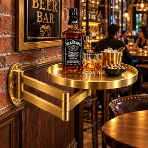 May include: A gold-coloured, wall-mounted, articulating swing-arm shelf with a circular tray. The shelf holds a bottle of Jack Daniel's Tennessee Whiskey, a glass filled with whiskey and ice, a gold ice bucket, and a small bowl of mixed nuts.