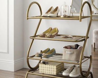 May include: A three-tier gold metal shoe rack with curved side frames. The shelves hold various items including white pumps, a glass cloche with jewellery, a small mirror, beige flats, a beige fabric storage bin, a plaid box, and beige trainers.
