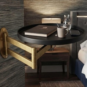 May include: A wall-mounted bedside table featuring a round black tray surface supported by an adjustable, articulated brass-toned metal arm. The tray holds a closed book, a ceramic mug, a glass carafe, and a smartphone, providing a compact surface.
