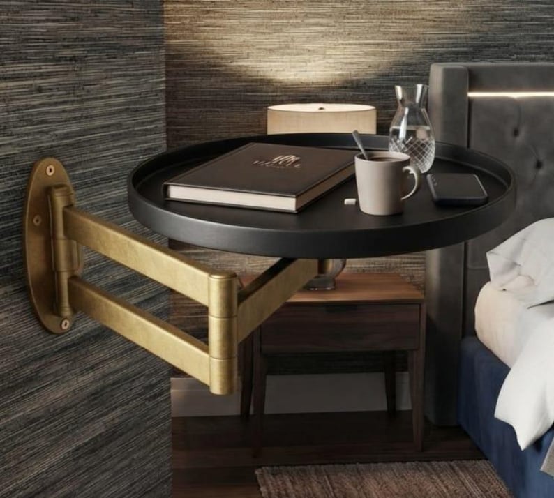 May include: A wall-mounted bedside table featuring a round black tray surface supported by an adjustable, articulated brass-toned metal arm. The tray holds a closed book, a ceramic mug, a glass carafe, and a smartphone, providing a compact surface.
