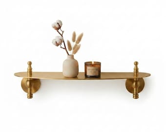 May include: A gold-toned metal wall shelf with two decorative finial supports. On the shelf sits a light-coloured ceramic vase containing dried cotton stems and beige grass plumes, alongside a brown glass candle jar with a visible label.