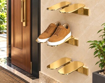 May include: Three pairs of wall-mounted gold-coloured metal shoe racks with a smooth, reflective finish. One pair of tan suede trainers with white rubber soles is placed on the middle rack, while the top and bottom racks remain empty.