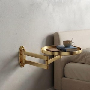 May include: A gold-toned metal wall-mounted swing arm side table with a circular tray top. The table features an adjustable articulated arm attached to a vertical wall plate. A ceramic mug and a closed book rest on the circular surface.