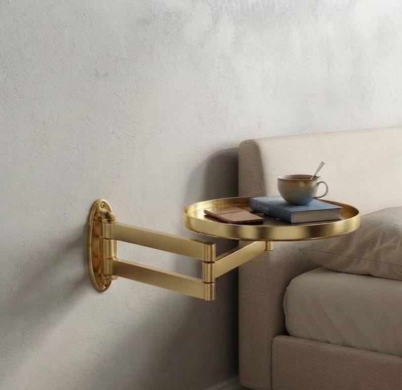 May include: A gold-toned metal wall-mounted swing arm side table with a circular tray top. The table features an adjustable articulated arm attached to a vertical wall plate. A ceramic mug and a closed book rest on the circular surface.