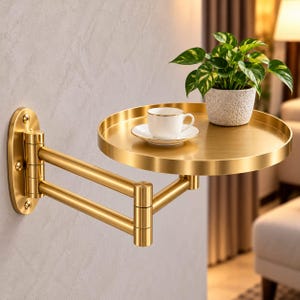 May include: A gold-toned metal wall-mounted shelf with a circular tray and an adjustable swing arm mechanism. A small white ceramic teacup and saucer sit on the tray, alongside a potted green plant in a textured light gray container.