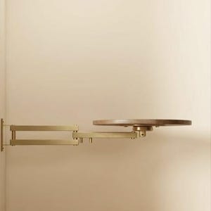 May include: A wall-mounted shelf featuring a matte gold-toned metal articulated swing arm and a circular wooden platform. The adjustable arm extends horizontally from the wall, supporting the round, flat wooden surface at its end.
