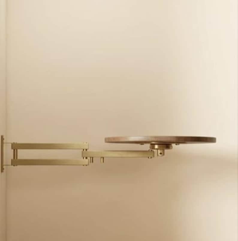 May include: A wall-mounted shelf featuring a matte gold-toned metal articulated swing arm and a circular wooden platform. The adjustable arm extends horizontally from the wall, supporting the round, flat wooden surface at its end.