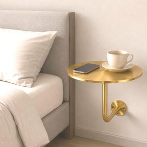 May include: A circular, gold-toned metal wall-mounted shelf featuring a single curved support arm attached to a circular base plate. A black smartphone and a beige ceramic mug on a matching saucer are placed on the flat, round surface.