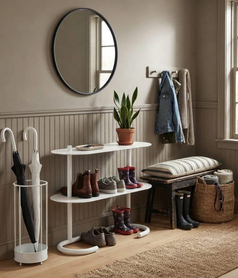 May include: A white two-tier oval console table holding a potted snake plant, brown leather boots, and grey trainers. Beside it, a white cylindrical umbrella stand holds two umbrellas, and a dark wooden bench features a striped cushion.