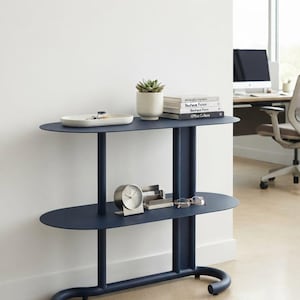 May include: A dark blue metal console table with two oval shelves and a curved base. The top shelf holds a white tray, a small potted succulent, and a stack of books. The bottom shelf features a silver analogue clock, a card holder, and glasses.