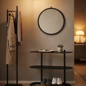 May include: A black metal coat rack with a beige trench coat and a scarf, a round black-framed wall mirror, and a black three-tiered oval console table holding a small succulent in a pot and a shallow dish with keys.