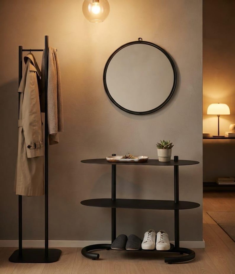 May include: A black metal coat rack with a beige trench coat and a scarf, a round black-framed wall mirror, and a black three-tiered oval console table holding a small succulent in a pot and a shallow dish with keys.