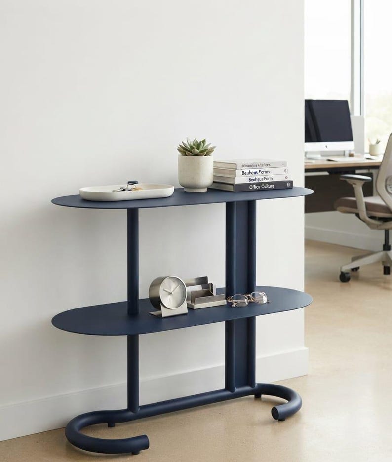 May include: A dark blue metal console table with two oval shelves and a curved tubular base. The top shelf holds a white tray, a small potted succulent, and a stack of books. The bottom shelf features a silver analogue clock and spectacles.